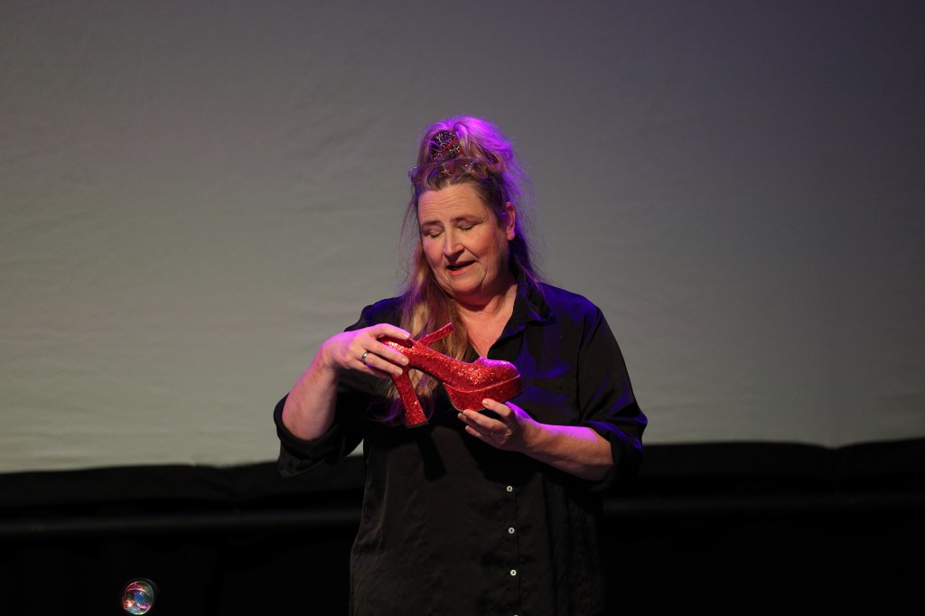 Cathy Crabb on stage holding a red glittery high-heeled shoe, lit by purple stage lighting
