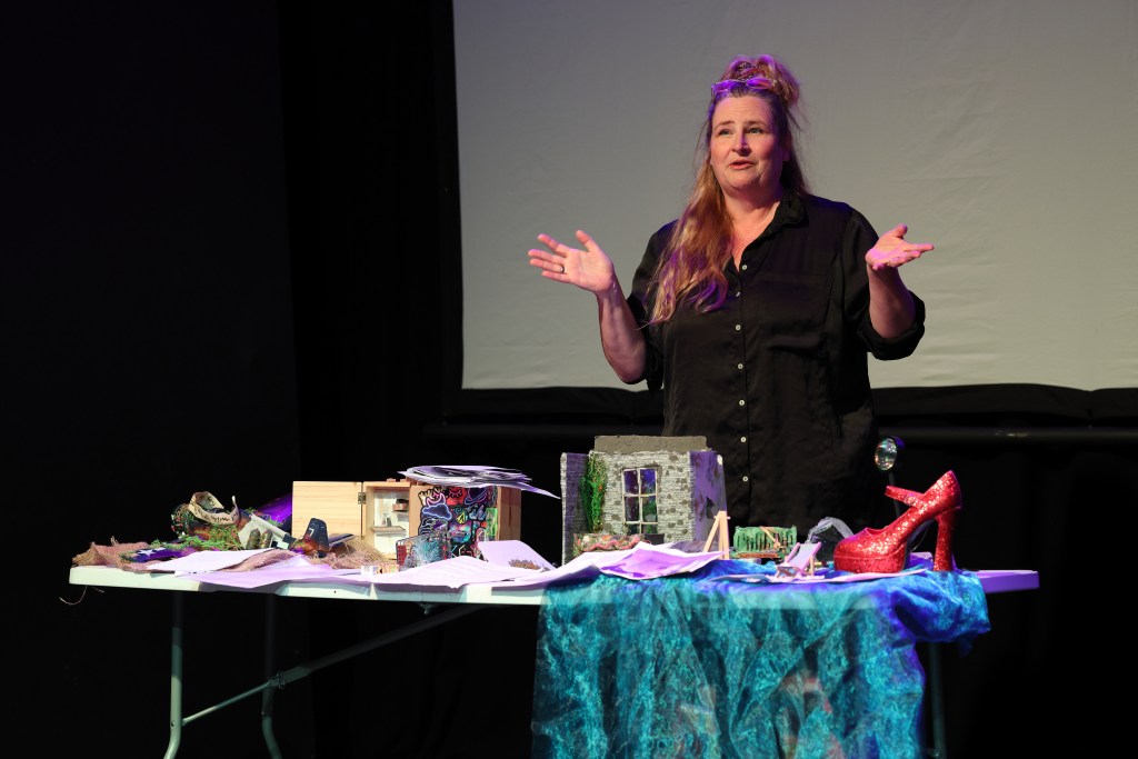 Cathy Crabb presenting on stage at a creative writing event, standing behind a table covered with colourful props including a miniature house, a red glittery shoe, fabric and handwritten notes