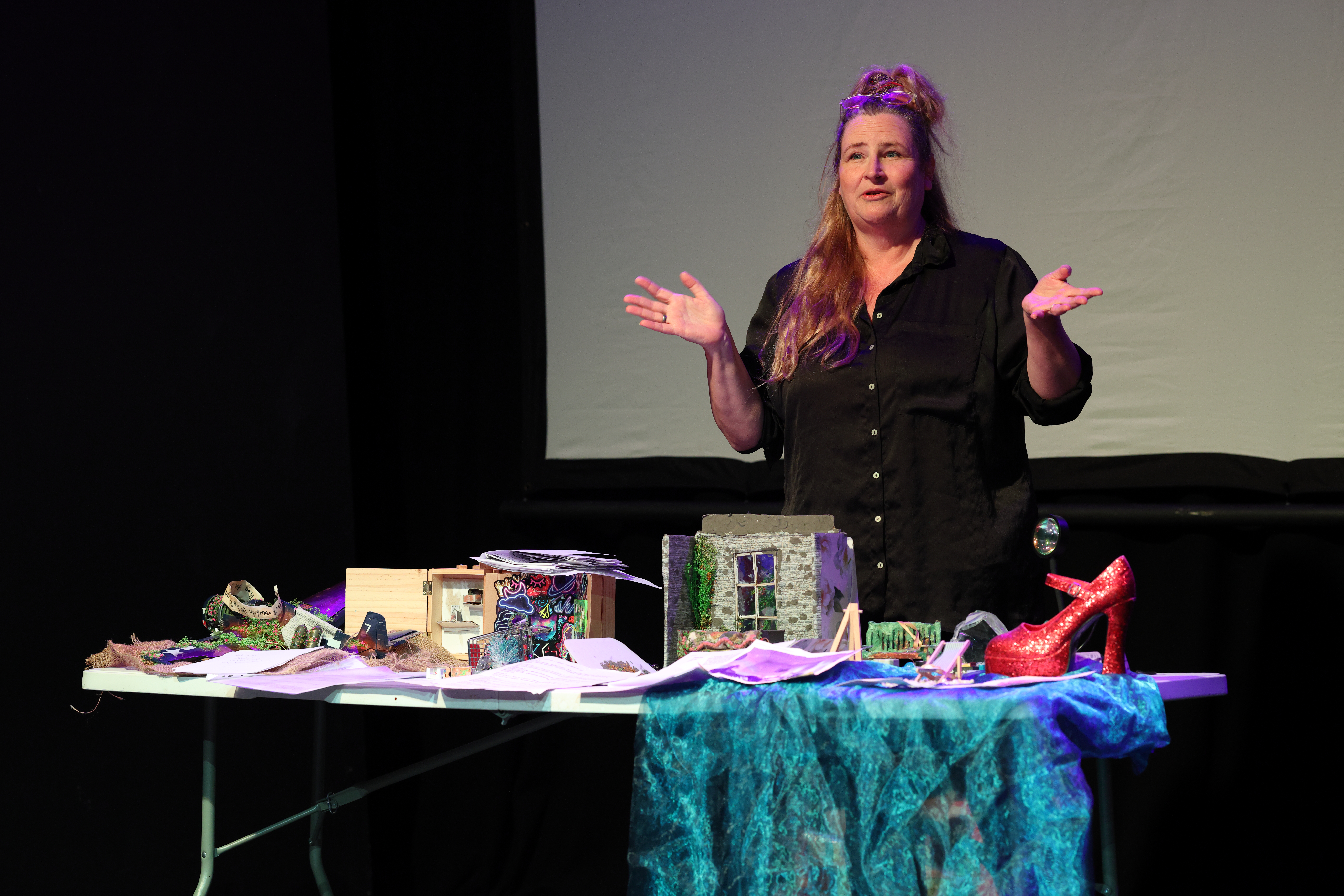 A performer stands behind a table of colorful theatrical props and set pieces, presenting on stage under purple and blue lighting.